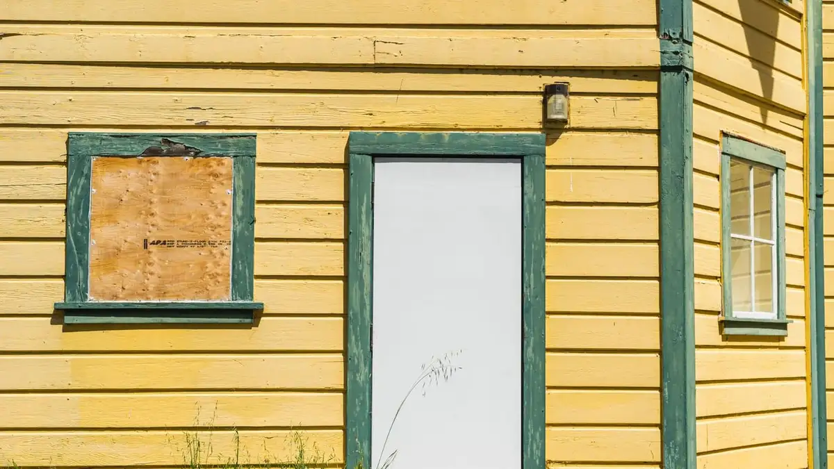 Window boarded up with plywood on a yellow timber house beside a white door