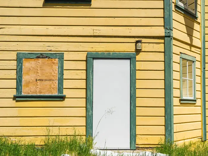 Window boarded up with plywood on a yellow timber house beside a white door