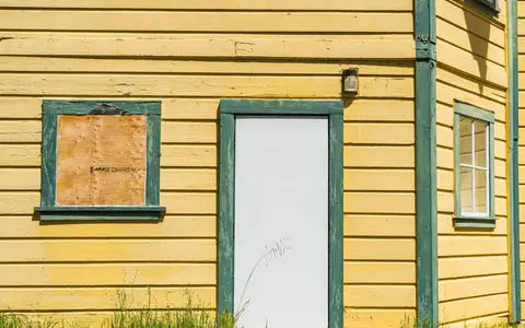 Window boarded up with plywood on a yellow timber house beside a white door