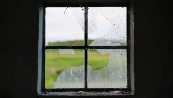 Wooden window frame with shattered glass panes, green fields beyond